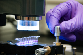 Researcher uses optical coherence tomography (OCT). A hand in a purple glove turns a metal dial on a plate holding tiny vials of purple liquid.